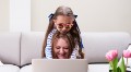 Portrait of smiling mother and daughter in sunglasses surfing the Internet via laptop against couch.